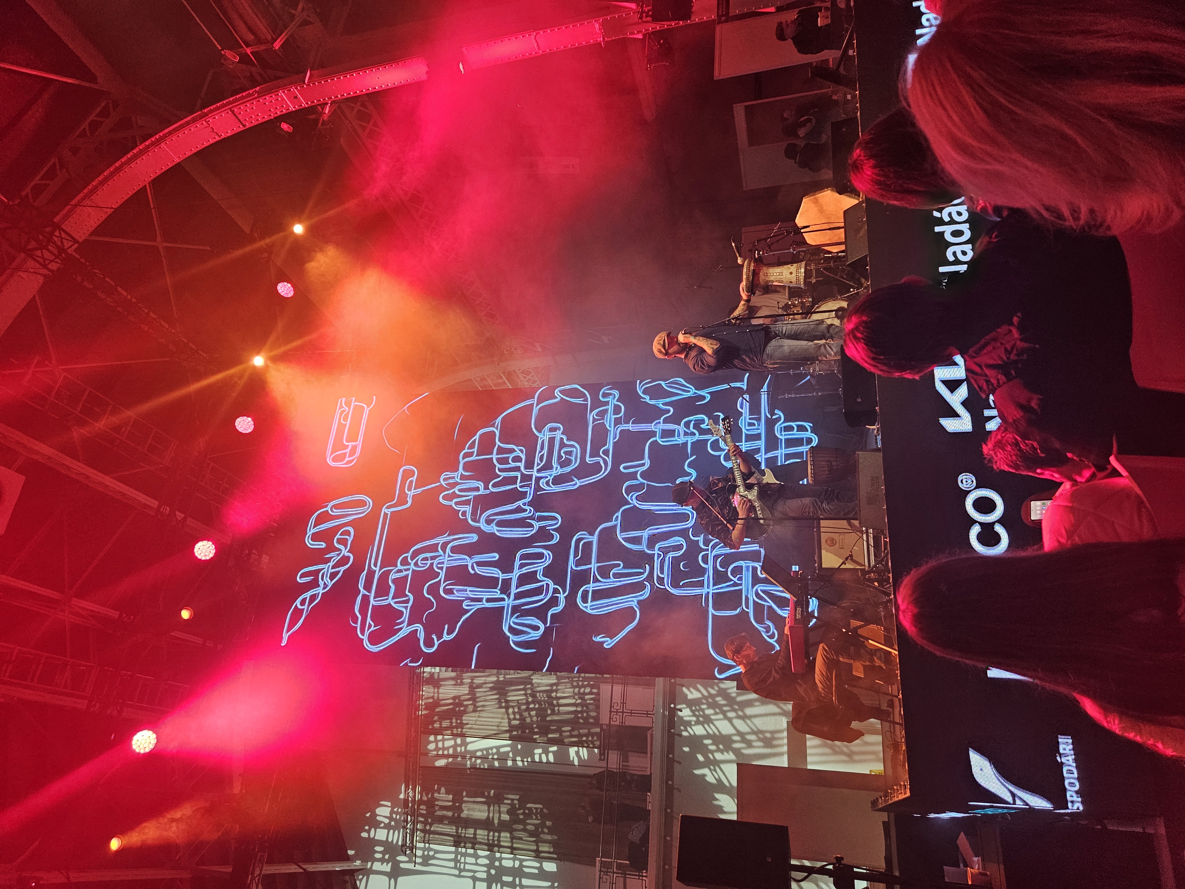 Koncert na żywo. Widać scenę oświetloną czerwonym światłem i sztuczną mgłą. Na scenie gra zespół muzyczny – wokalista stoi na środku, obok niego gitarzysta, a inny muzyk gra na instrumencie klawiszowym. W tle znajduje się duży ekran z abstrakcyjną grafiką w stylu neonowym, przedstawiającą nieregularne kształty.