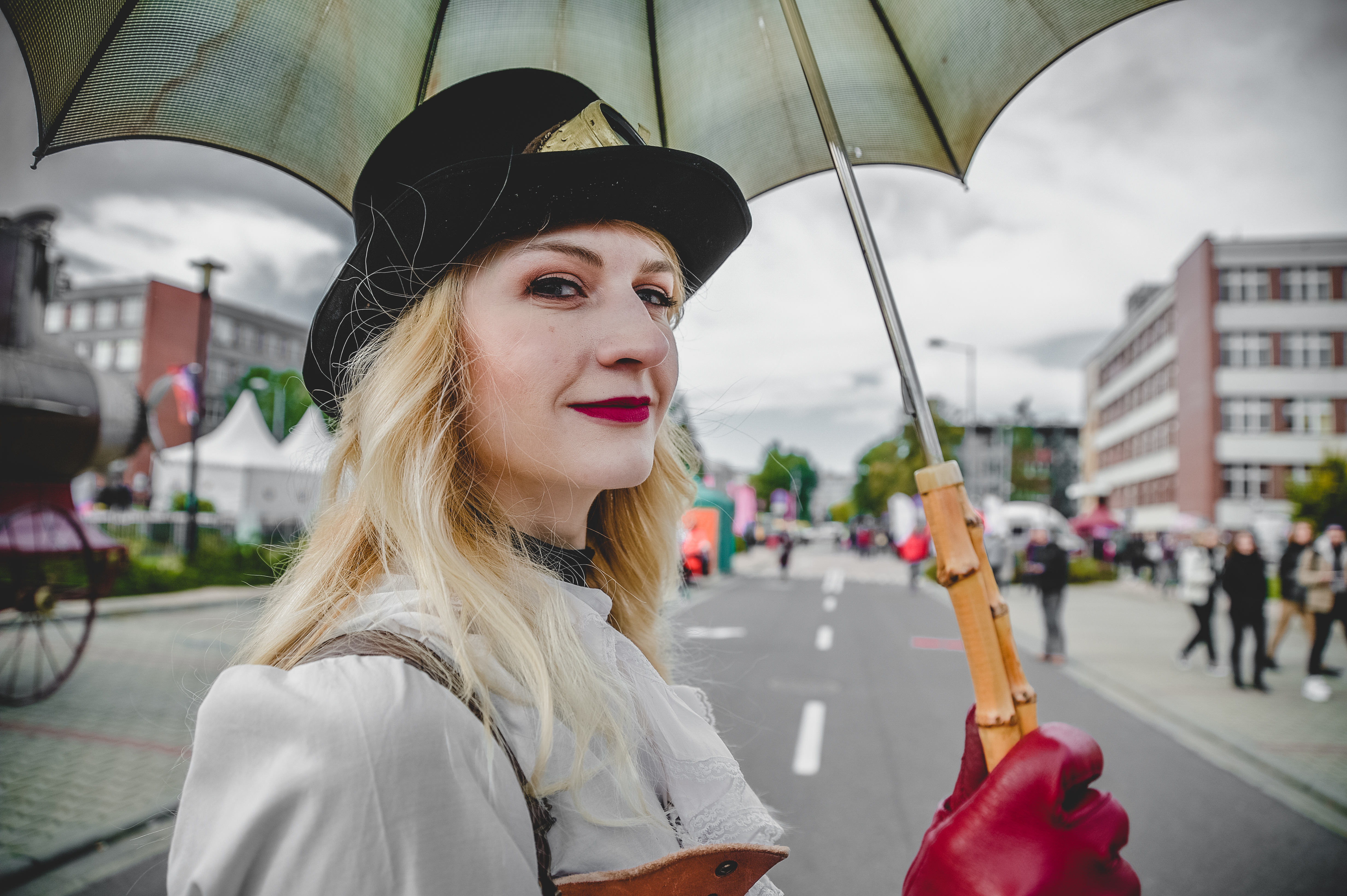 Kobieta w czarno-biało-czerwonym stroju steampunk trzyma retro parasol i spogląda w kamerę, w tle budynki wydziałów UŚ | fot. Wojtek Korpusik